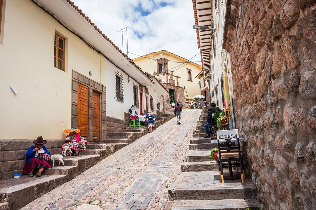 Vista diurna de una calle cusqueña (Calle Cuesta del Almirante), con varias personas interactuando y la luz del sol iluminando todo el ambiente